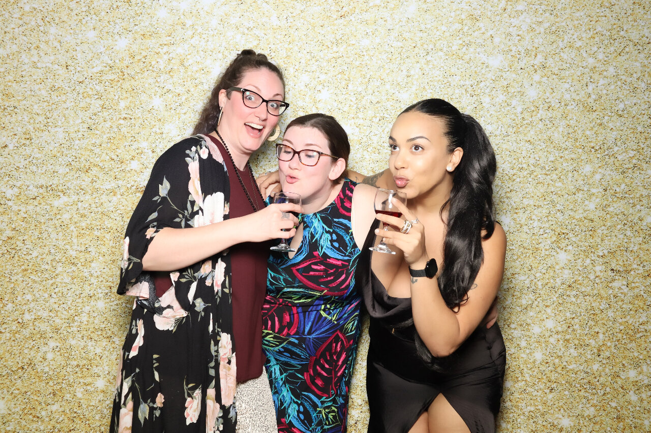 hree women stand in front of a glittery gold backdrop, holding wine glasses and making playful faces at the camera. They are dressed in evening attire and appear to be enjoying Evening of Elegance